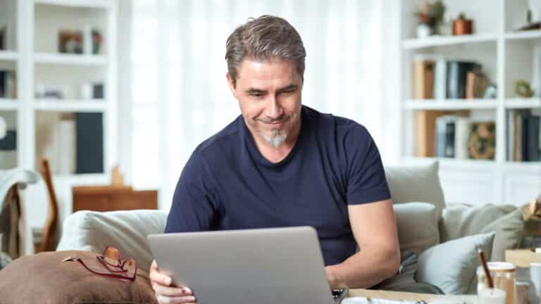 Mann sitzt lächelnd auf dem Sofa mit Laptop und informiert sich zur privaten Krankenversicherung (PKV).